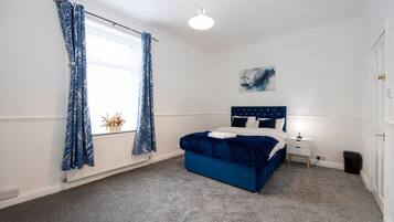 A bedroom with a bed, a bedside table, a lamp, a window with curtains, a radiator, and a wall-mounted artwork.