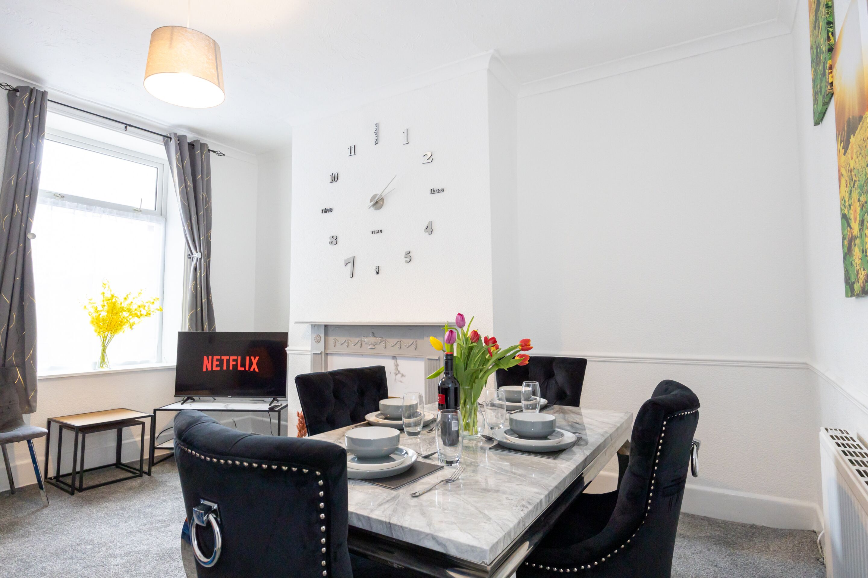 A modern dining area with a marble table, black upholstered chairs, a wall clock, and a flat-screen TV displaying 'NETFLIX'.