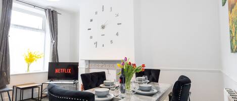 A modern dining area with a marble table, black upholstered chairs, a wall clock, and a flat-screen TV displaying 'NETFLIX'.