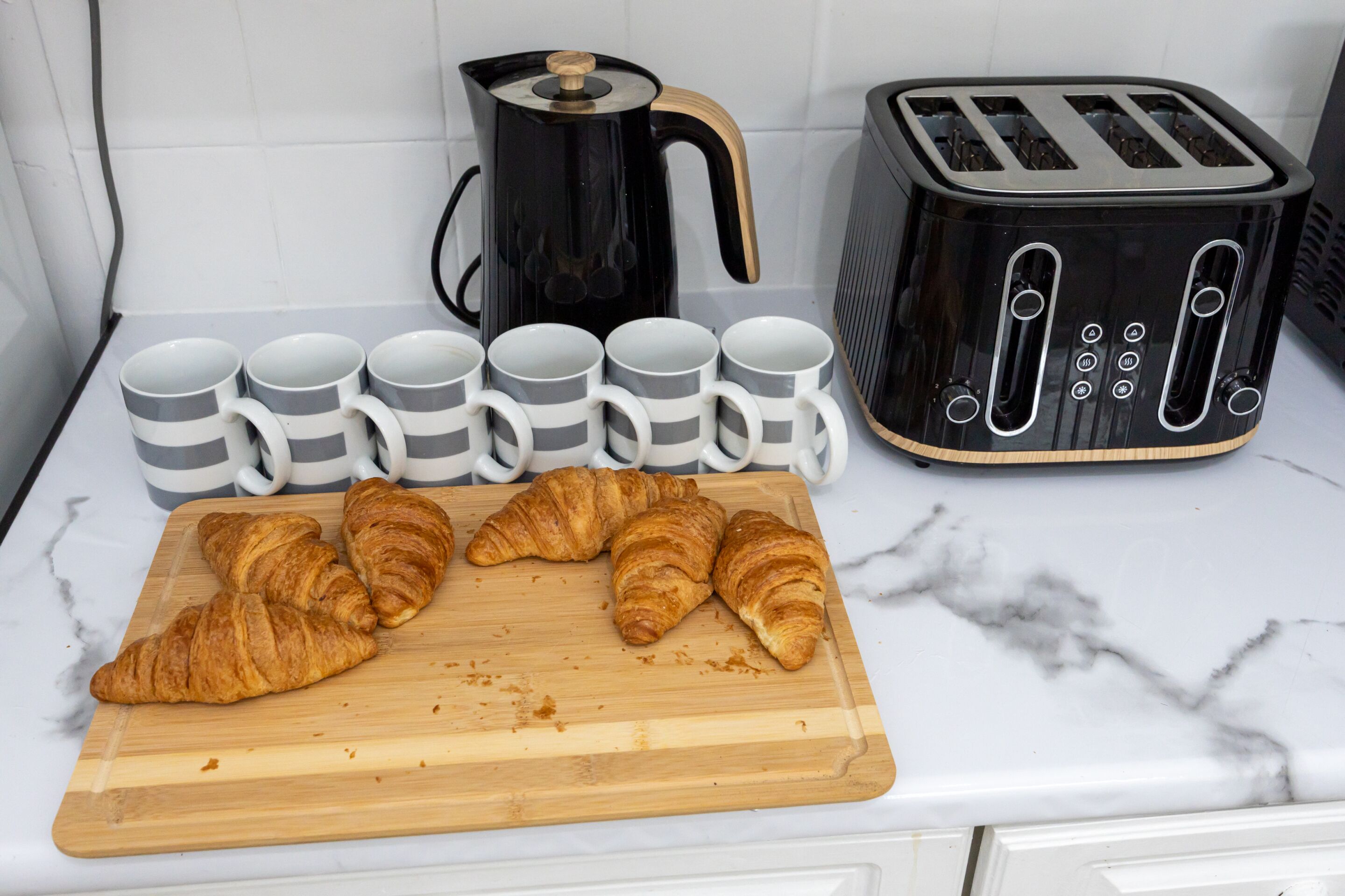 Een keukentafel met een broodrooster, een koffiezetapparaat en een houten snijplank met croissants.