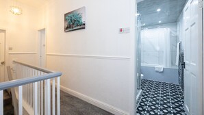 Een gang met een witte muur, een schilderij van bomen, een rookmelder en een glazen deur die leidt naar een badkamer met een zwart-wit geruit vloertje.