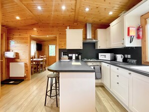 A wooden cabin interior with a kitchen featuring white cabinets, a black countertop, and a central island with stools. - Magnolia Lodge-UK46280 (Camerton, near Bath)