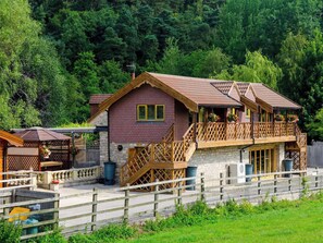 A two-story wooden cabin with a balcony, surrounded by a fence and lush greenery.