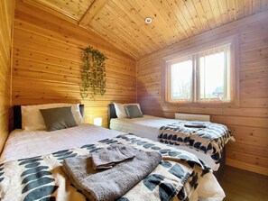 A cabin room with two beds, wooden walls, a hanging plant, and a window with blinds.