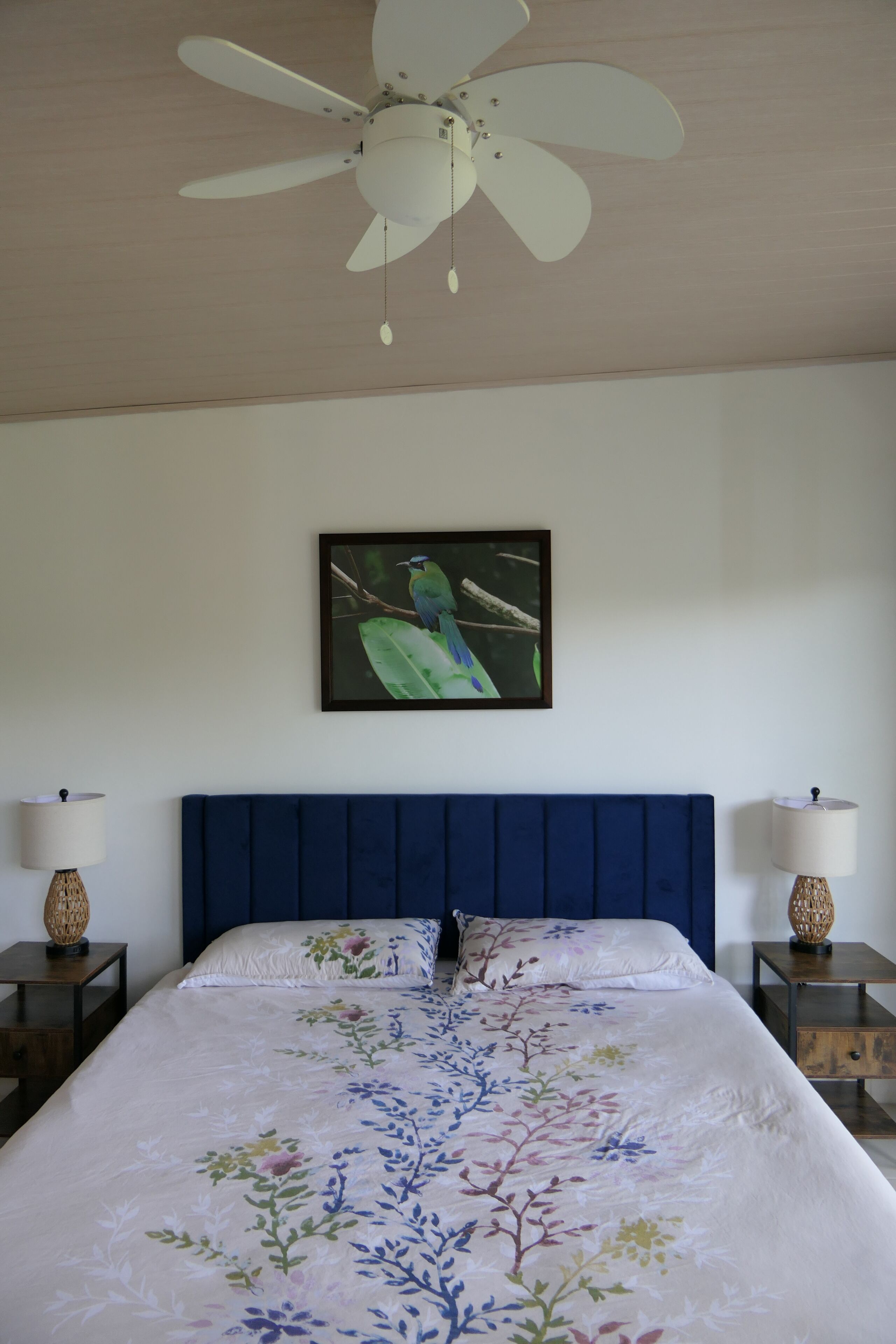 Un dormitorio con una cama grande, un ventilador de techo, un cuadro enmarcado de un pájaro y dos mesitas de noche con lámparas.