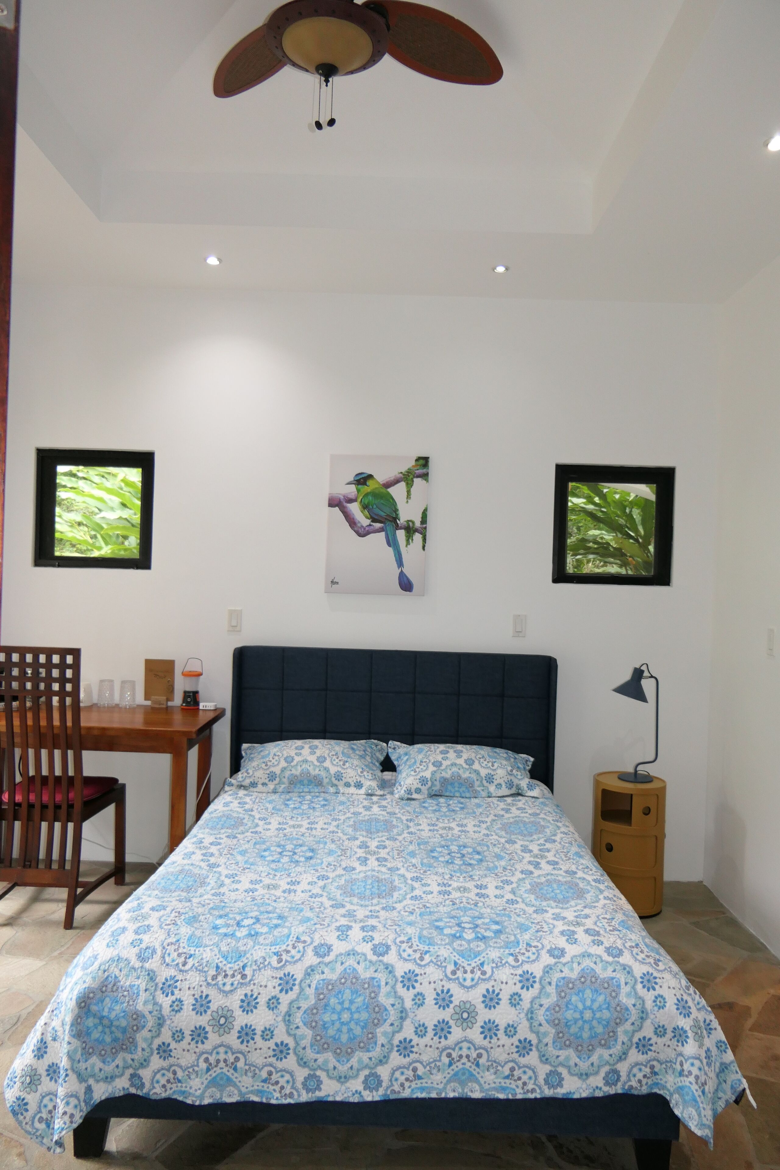 Un dormitorio con cama, ventilador de techo, escritorio con silla y dos cuadros en la pared.