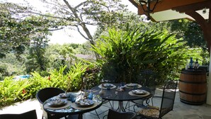 Une terrasse extérieure avec des tables et des chaises rondes, entourée d’une végétation luxuriante et d’une barrique en bois.