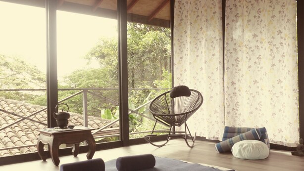 Una habitación con un ventanal grande, techo de madera, una mecedora, una pequeña mesa con tetera y una cama con almohadas.
