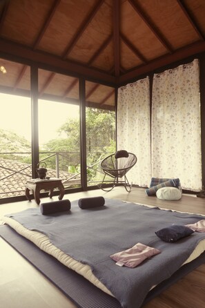 A room with a large window, a wooden ceiling, a rocking chair, a small table with a teapot, and a bed with pillows.