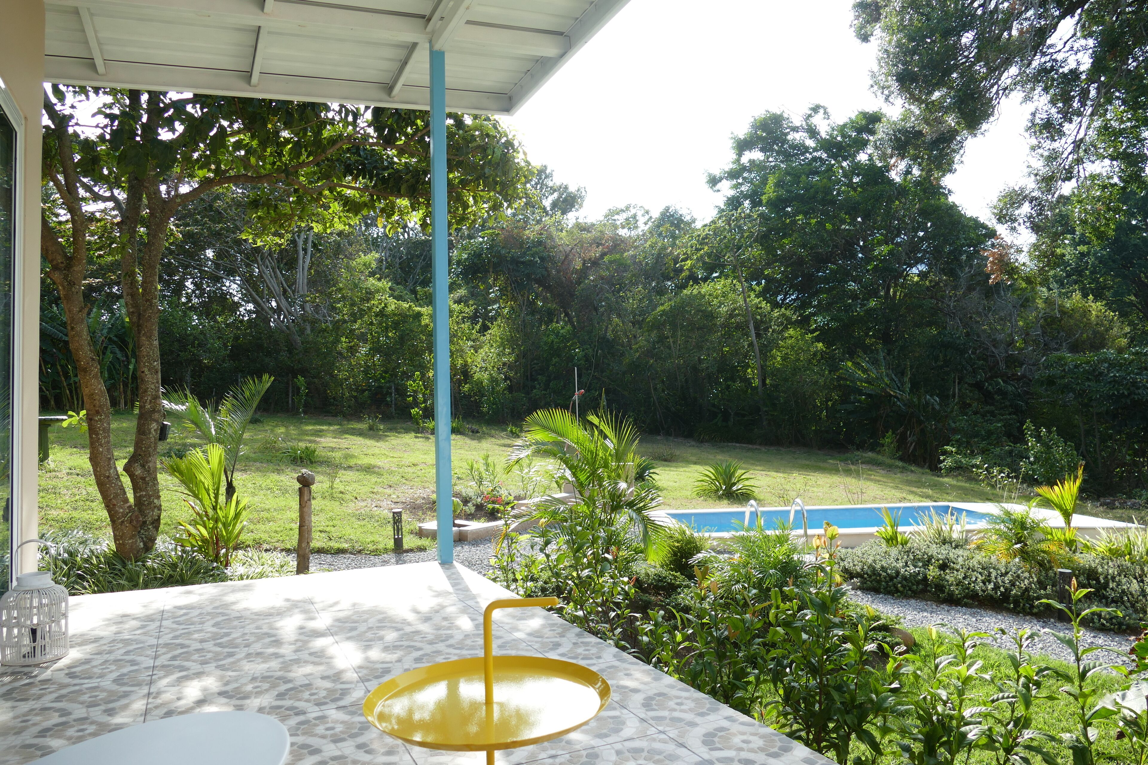 Un patio con una mesa amarilla y una mesa blanca, rodeado de exuberante follaje y una piscina.