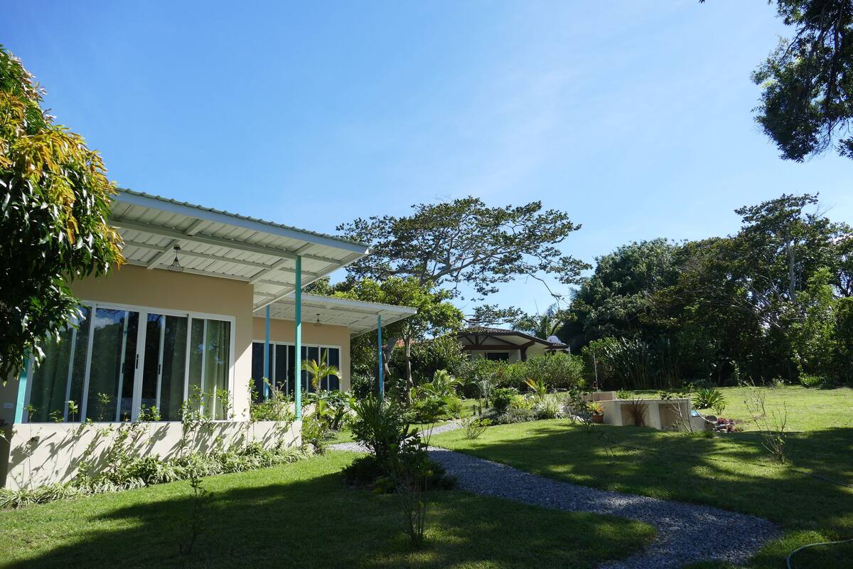 Una casa moderna con porche cubierto, rodeada de un jardín cuidado y árboles.