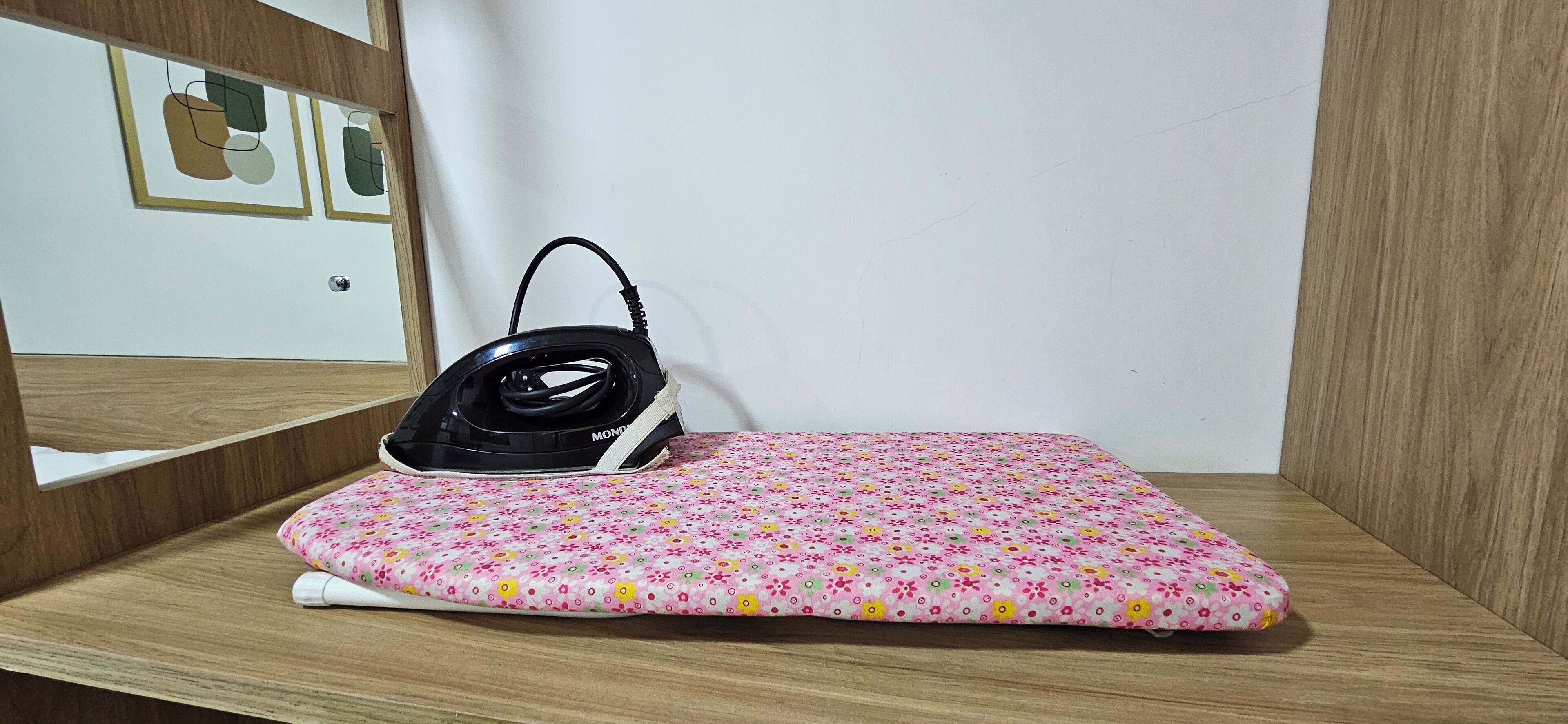 Un lit recouvert d’une couvre-lit à motifs floraux roses, avec un fer à repasser noir posé dessus.