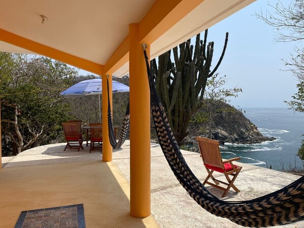 A covered outdoor seating area with a hammock, chairs, and a view of the ocean.