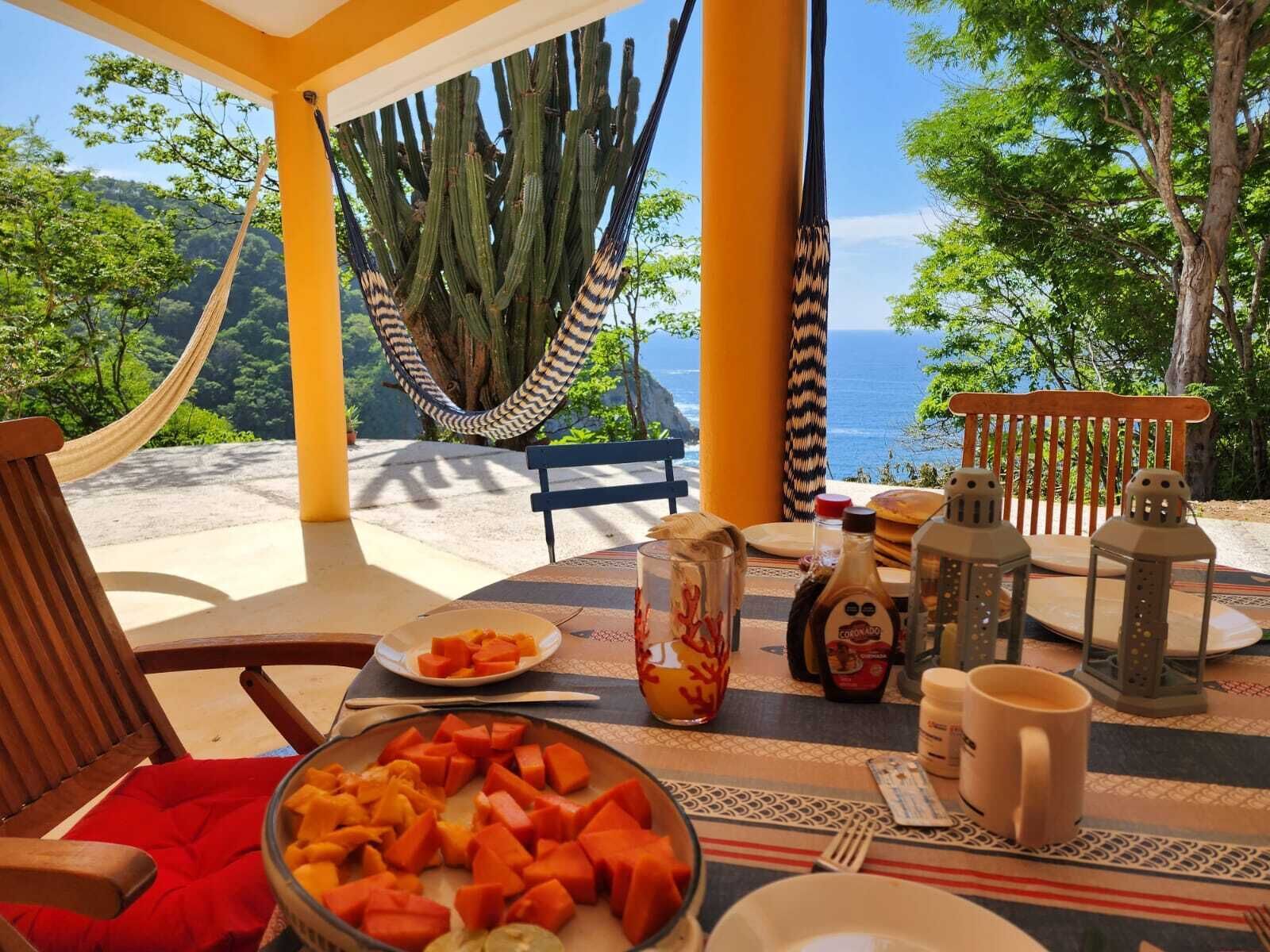 A dining area with a table set for two, a hammock, and a view of the sea and greenery.