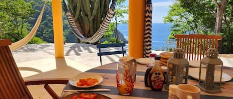 A dining area with a table set for two, a hammock, and a view of the sea and greenery.