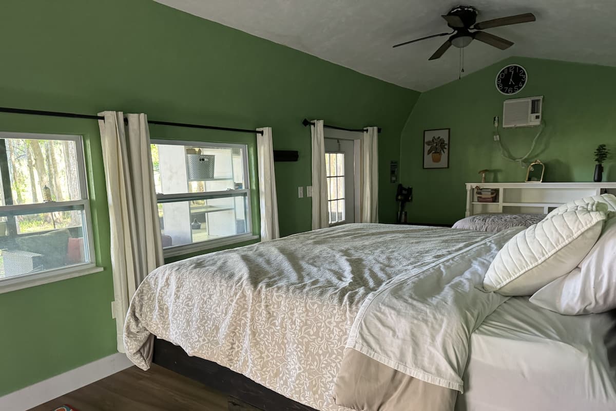 A bedroom with a large bed, a ceiling fan, a window with curtains, and a view of the outdoors.