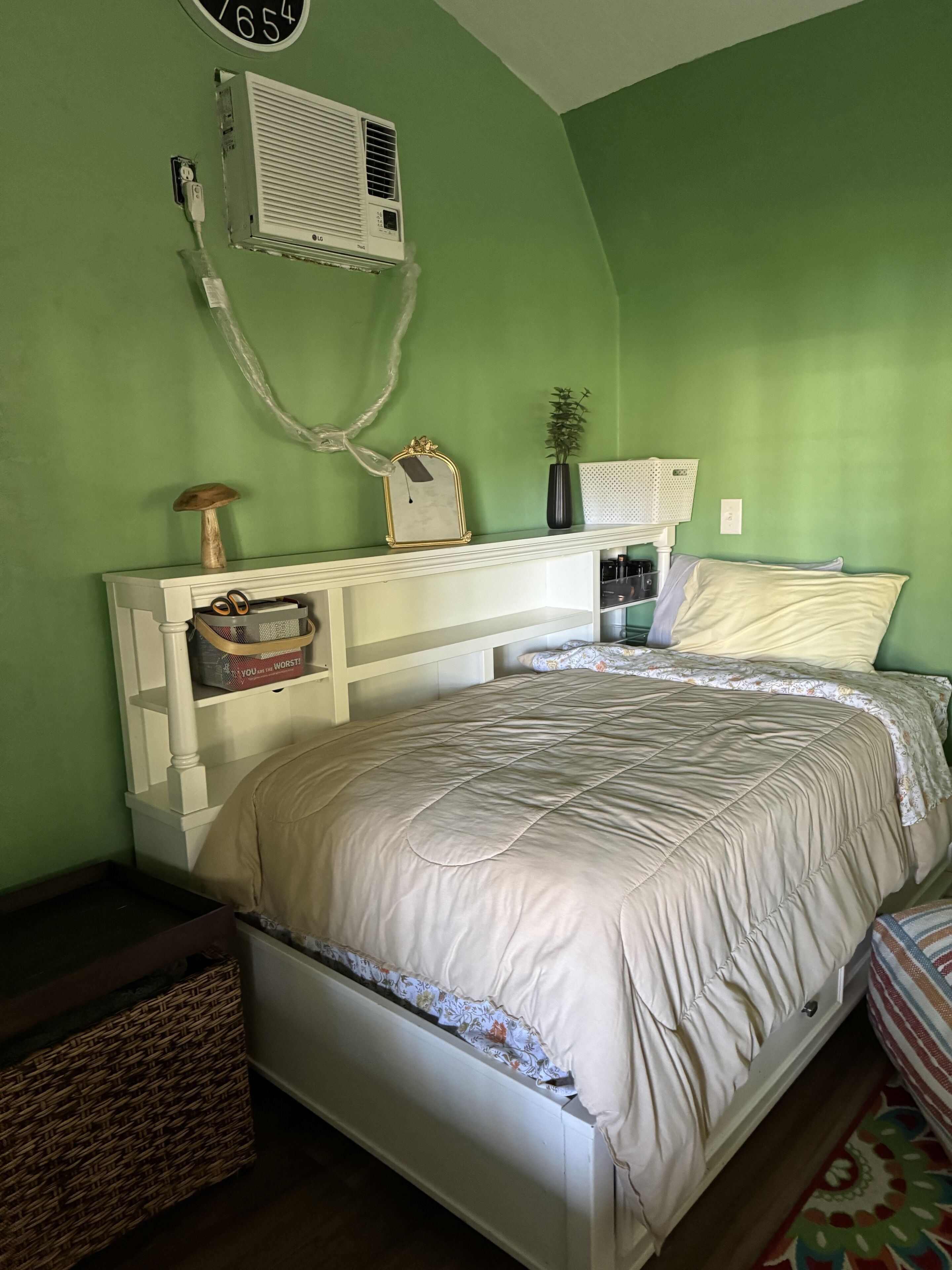 A bedroom with a white bed, green walls, a white headboard, a small shelf with a vase and a picture frame, and a white air conditioning unit on the wall.
