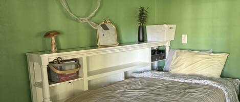 A bedroom with a white bed, green walls, a white headboard, a small shelf with a vase and a picture frame, and a white air conditioning unit on the wall.