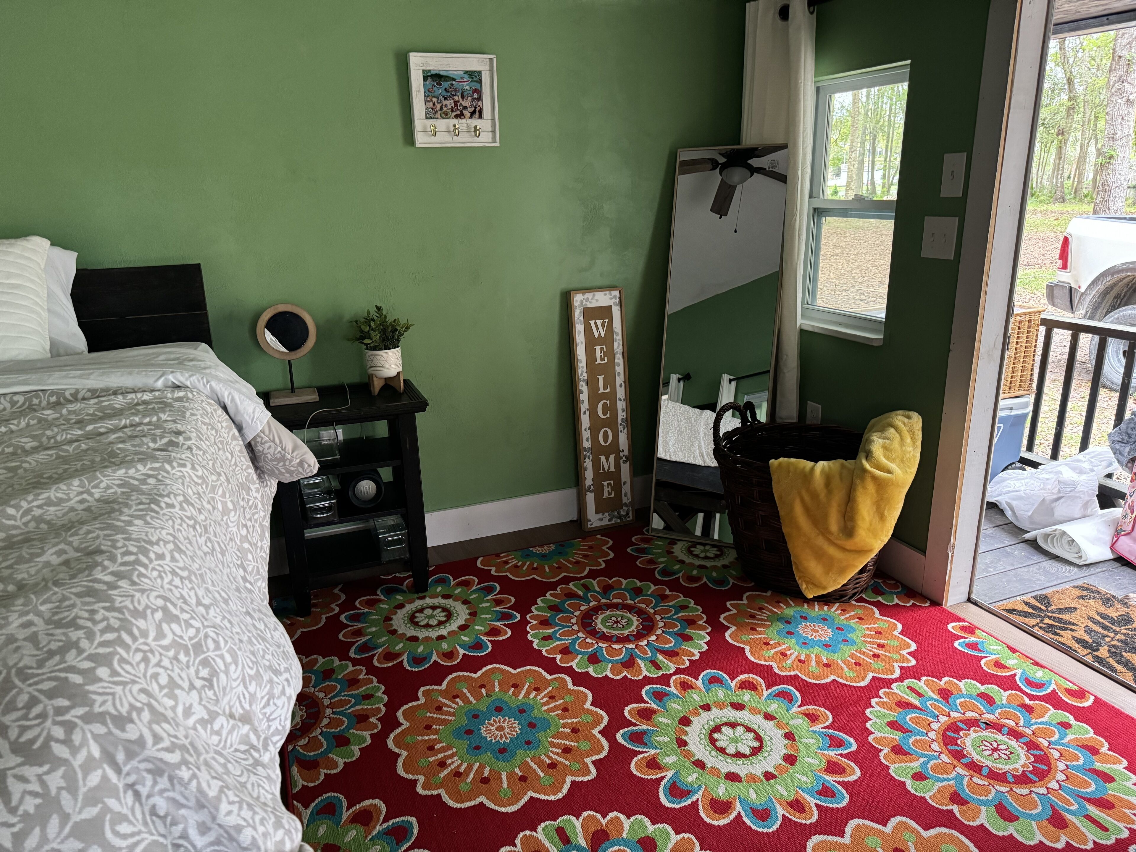 A bedroom with a bed, a bedside table with a plant, a mirror, and a colorful carpet.