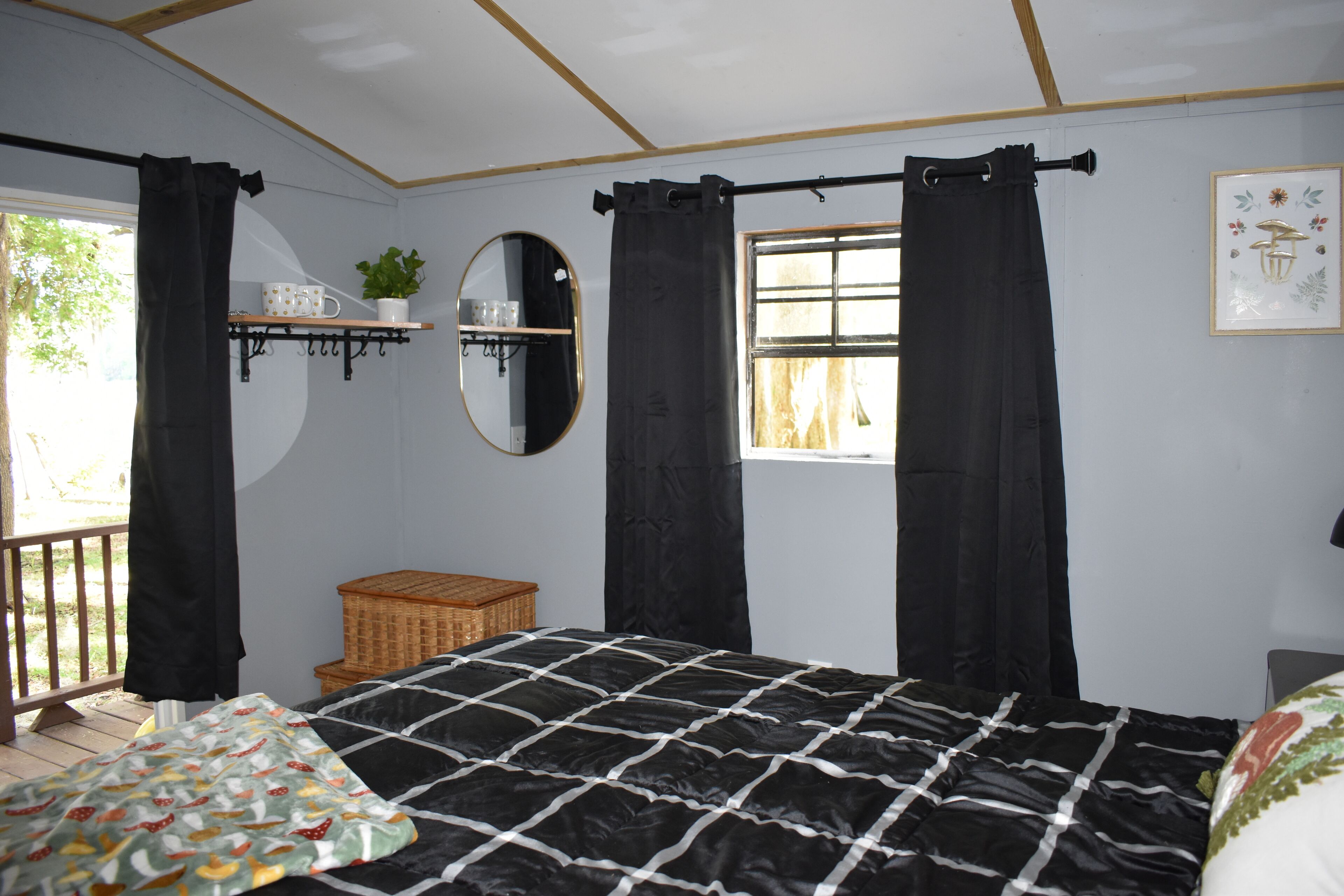 Ein Schlafzimmer mit einem großen Bett, schwarzen Vorhängen, einem Fenster, einem Spiegel und einem kleinen Regal mit einer Pflanze.