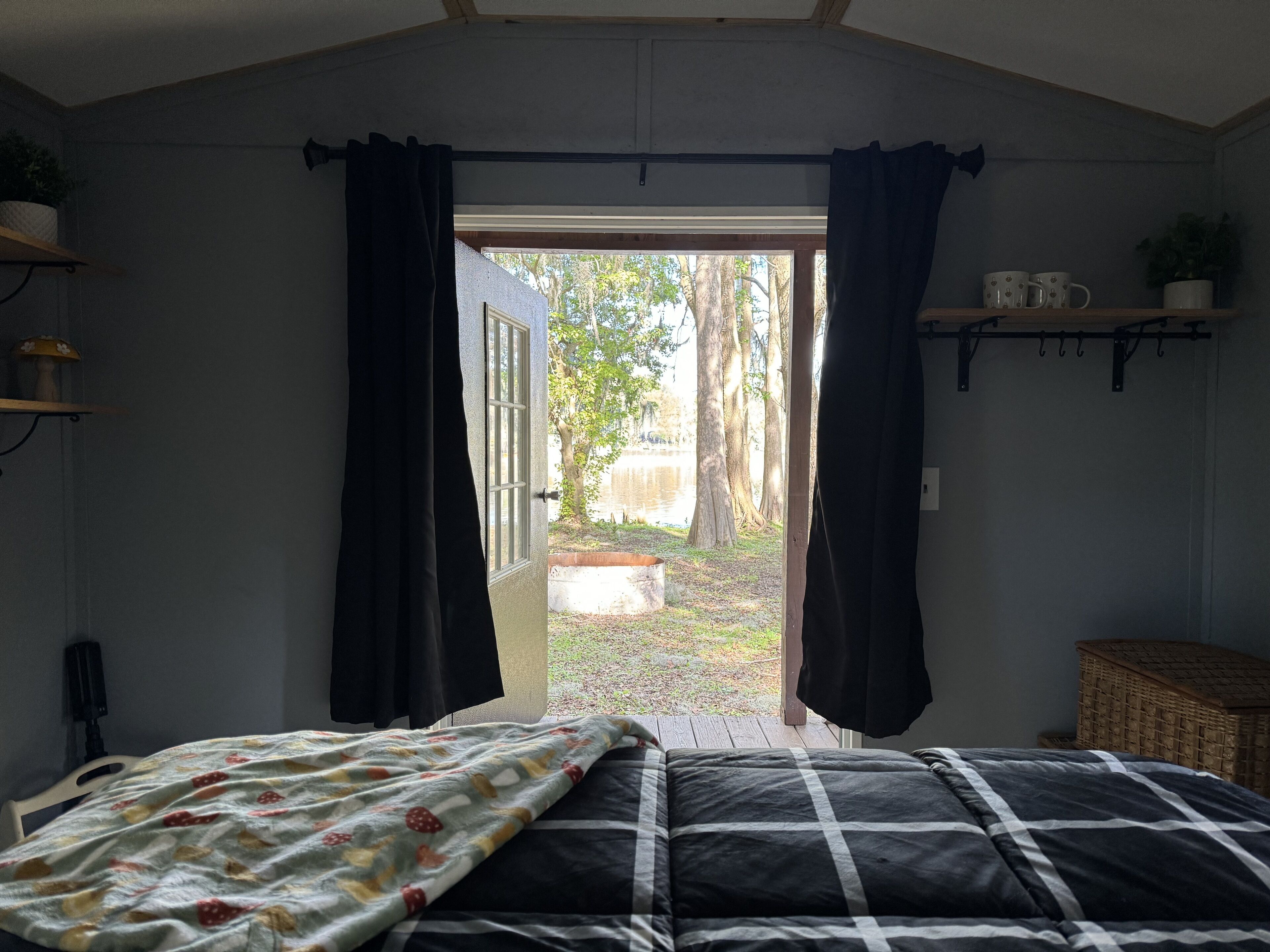 A bedroom with a bed, a window with curtains, a shelf with mugs, and a view of the outdoors.
