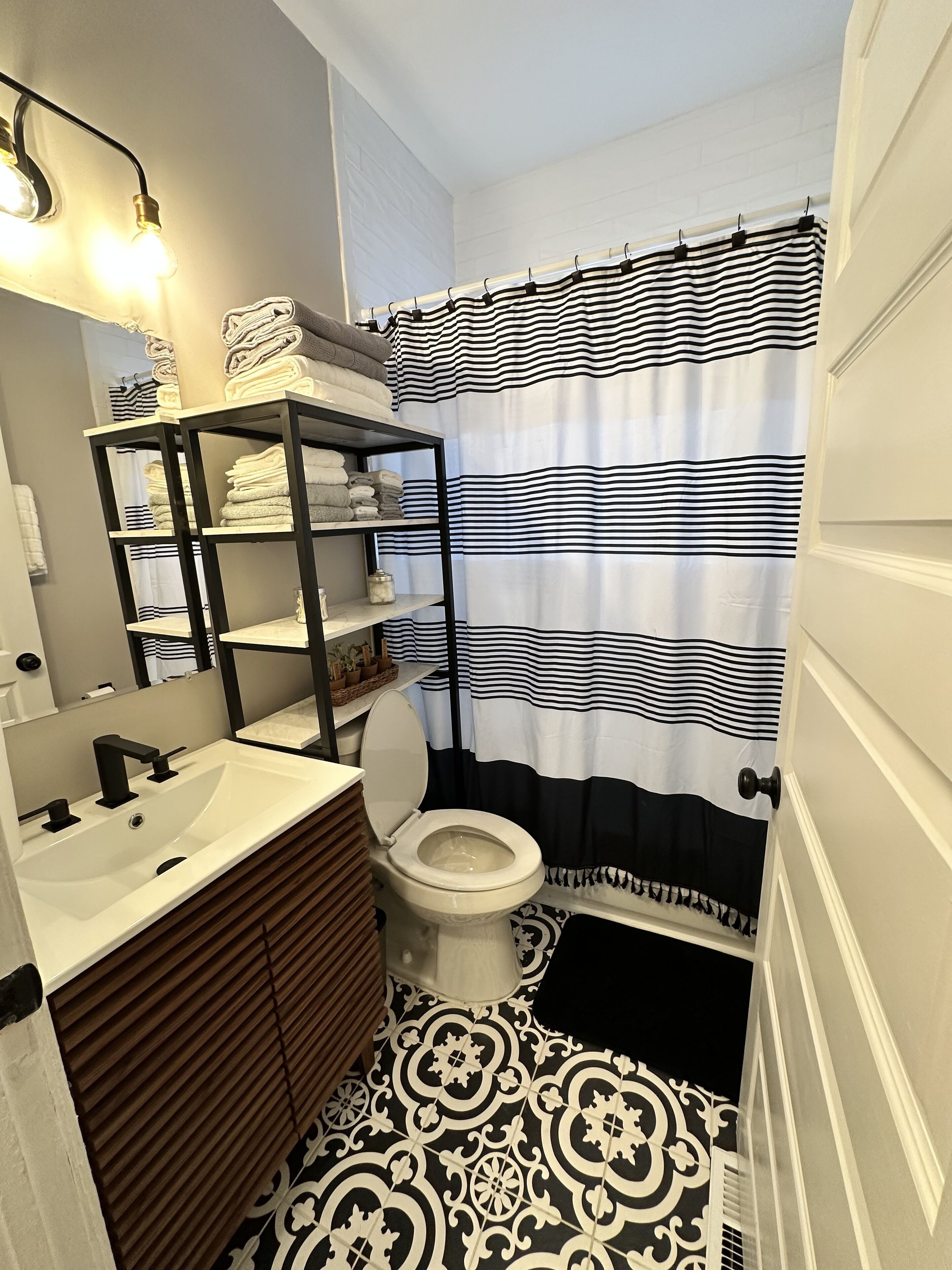 A bathroom with a white sink, black faucet, and a toilet. There is a shelf with towels and a mirror above the sink. The floor has a black and white patterned design.