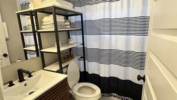A bathroom with a white sink, black faucet, and a toilet. There is a shelf with towels and a mirror above the sink. The floor has a black and white patterned design.