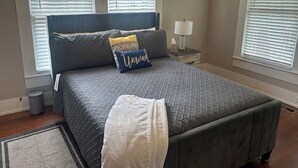 Un dormitorio con cama, mesitas de noche, lámpara y ventanas con persianas.