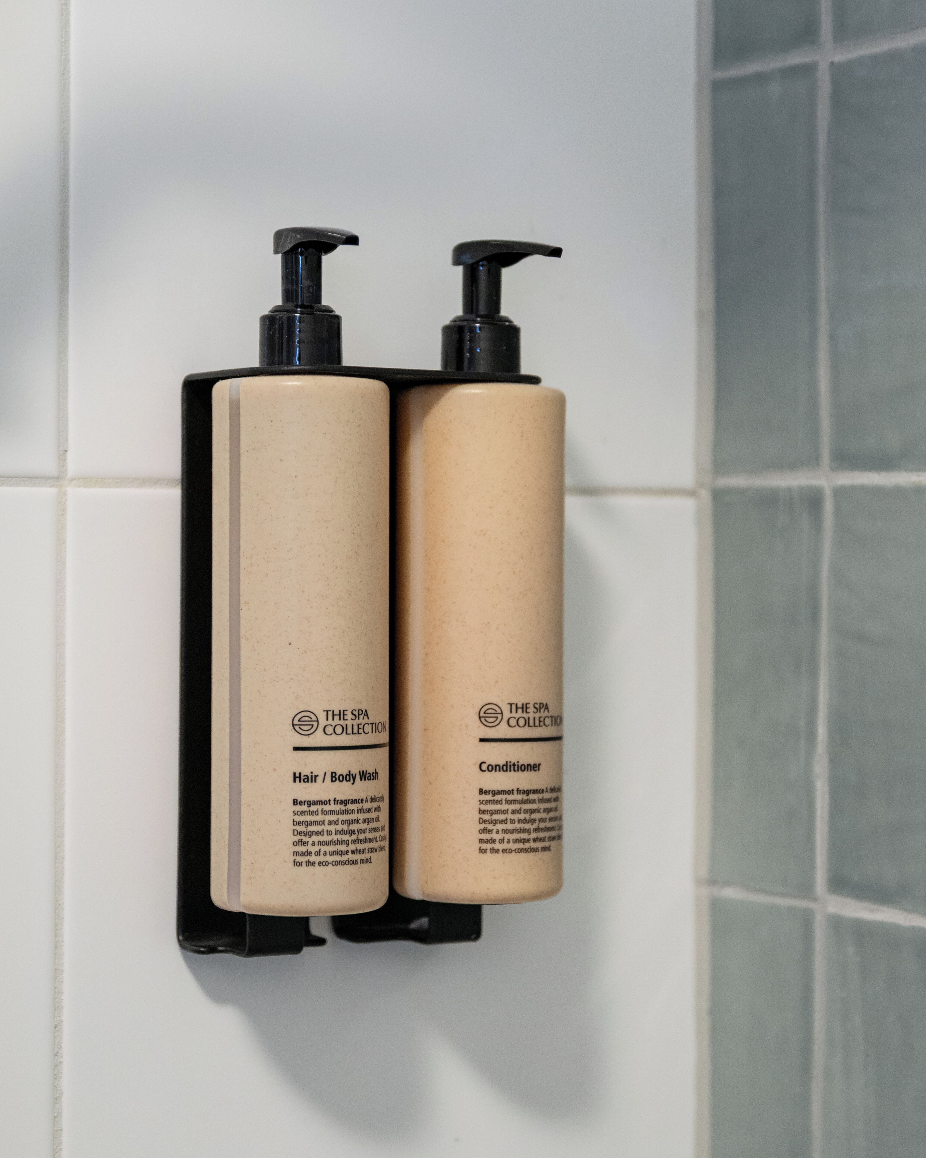 Dos botellas de la colección de spa, una con la etiqueta 'Champú y Gel de Ducha' y la otra 'Acondicionador', montadas en una pared de azulejos.