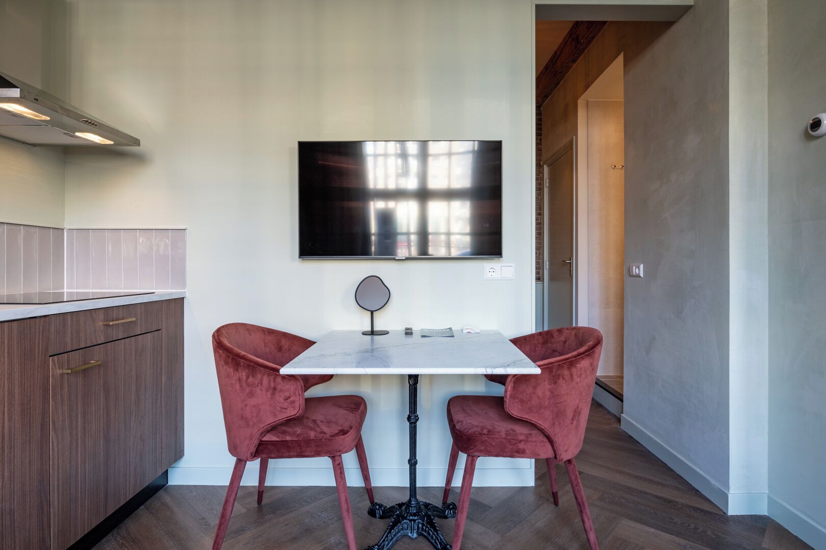 A modern kitchen with a marble countertop, two red velvet chairs, a small table, and a large flat-screen TV mounted on the wall.