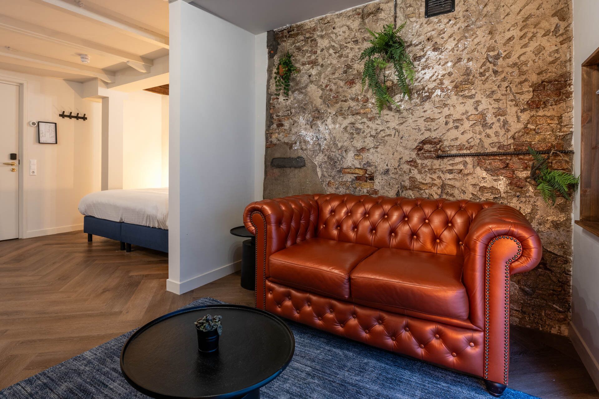 A room with a brown leather sofa, a bed, a stone wall, and a small table with a plant.
