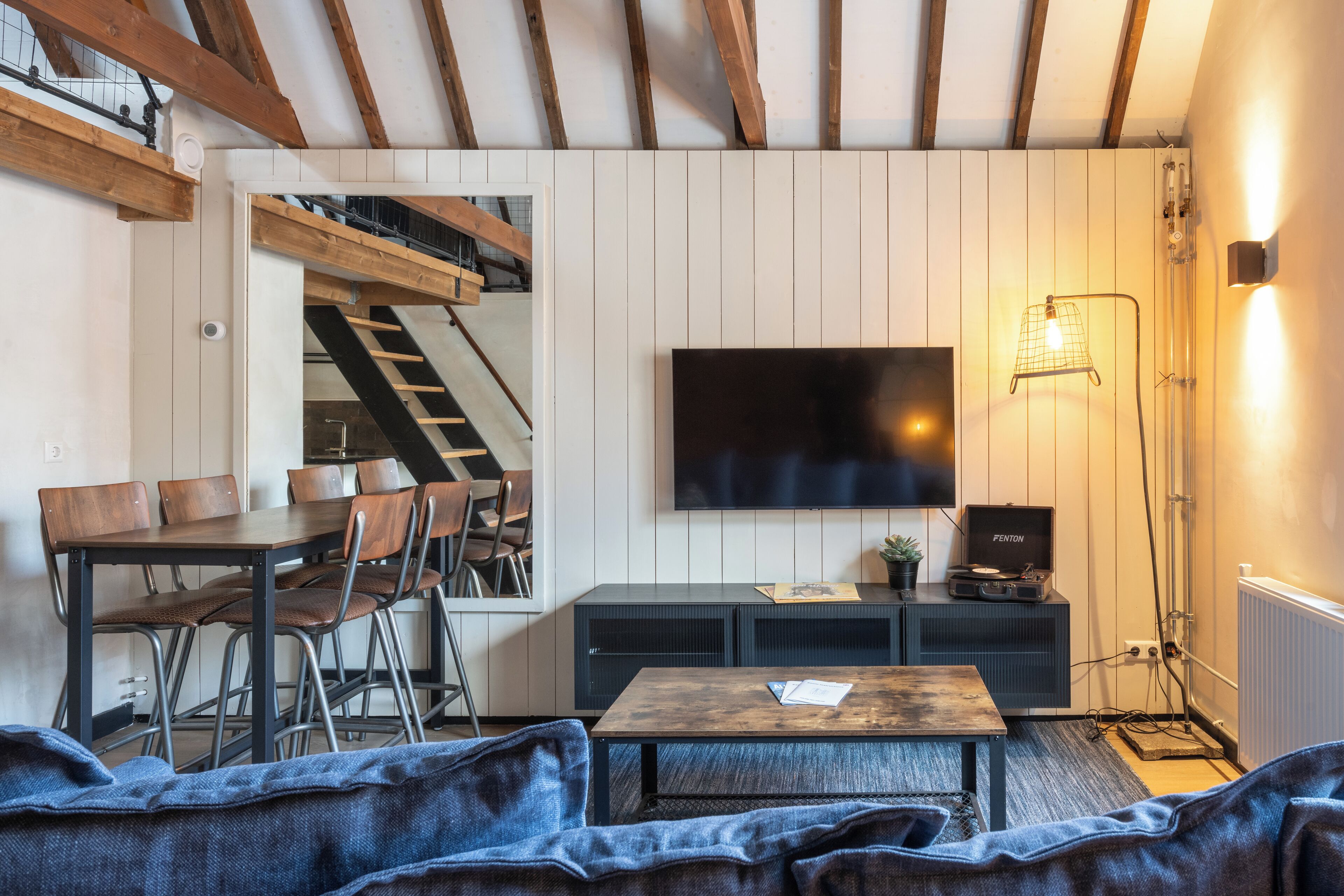 A cozy living room with a wooden staircase, a dining area, and a flat-screen TV mounted on the wall.