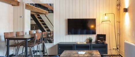 A cozy living room with a wooden staircase, a dining area, and a flat-screen TV mounted on the wall.