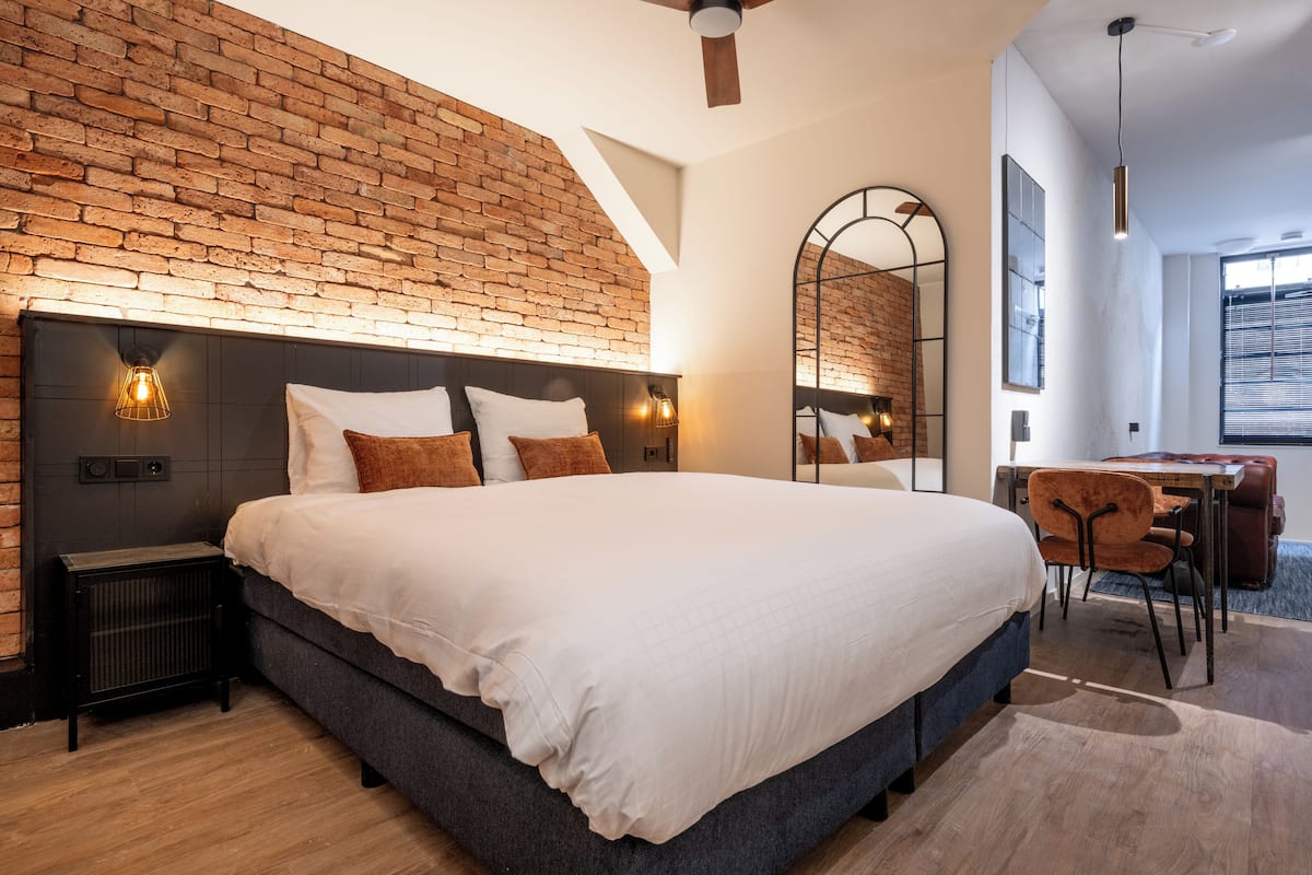 A modern bedroom with a brick wall, a large bed with white bedding, a desk with a chair, and a large mirror.