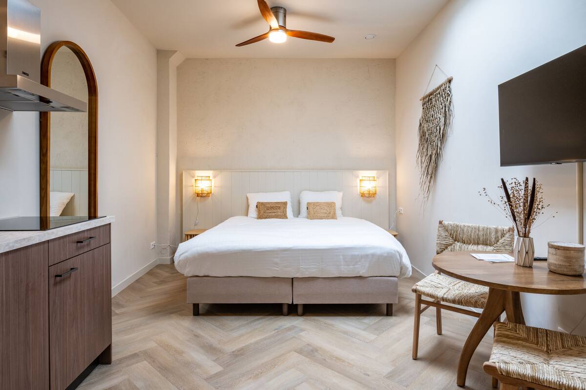 A modern bedroom with a large bed, bedside lamps, a ceiling fan, a TV, and a small dining area with a table and chairs.