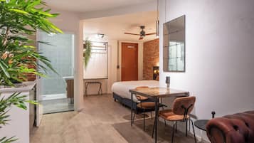 A compact hotel room with a bed, a small dining table, chairs, a mirror, a ceiling fan, and a potted plant.