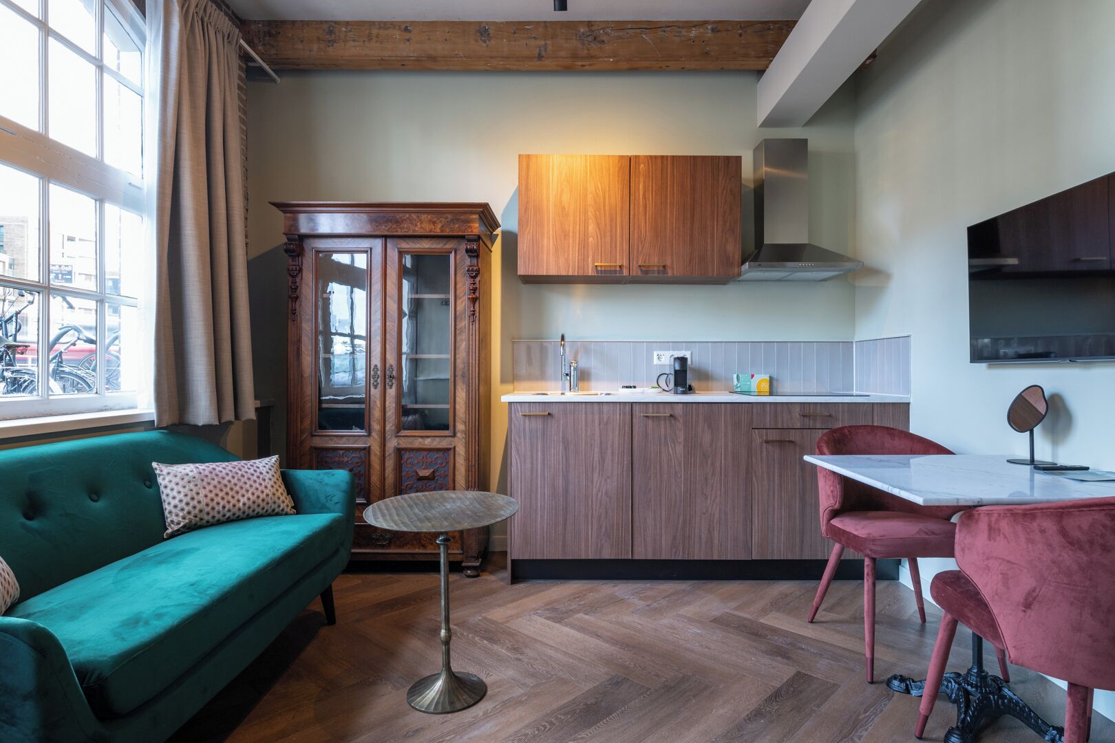 A compact living space with a teal sofa, a small round table, a wooden cabinet, and a kitchen area with a countertop and a TV mounted on the wall.