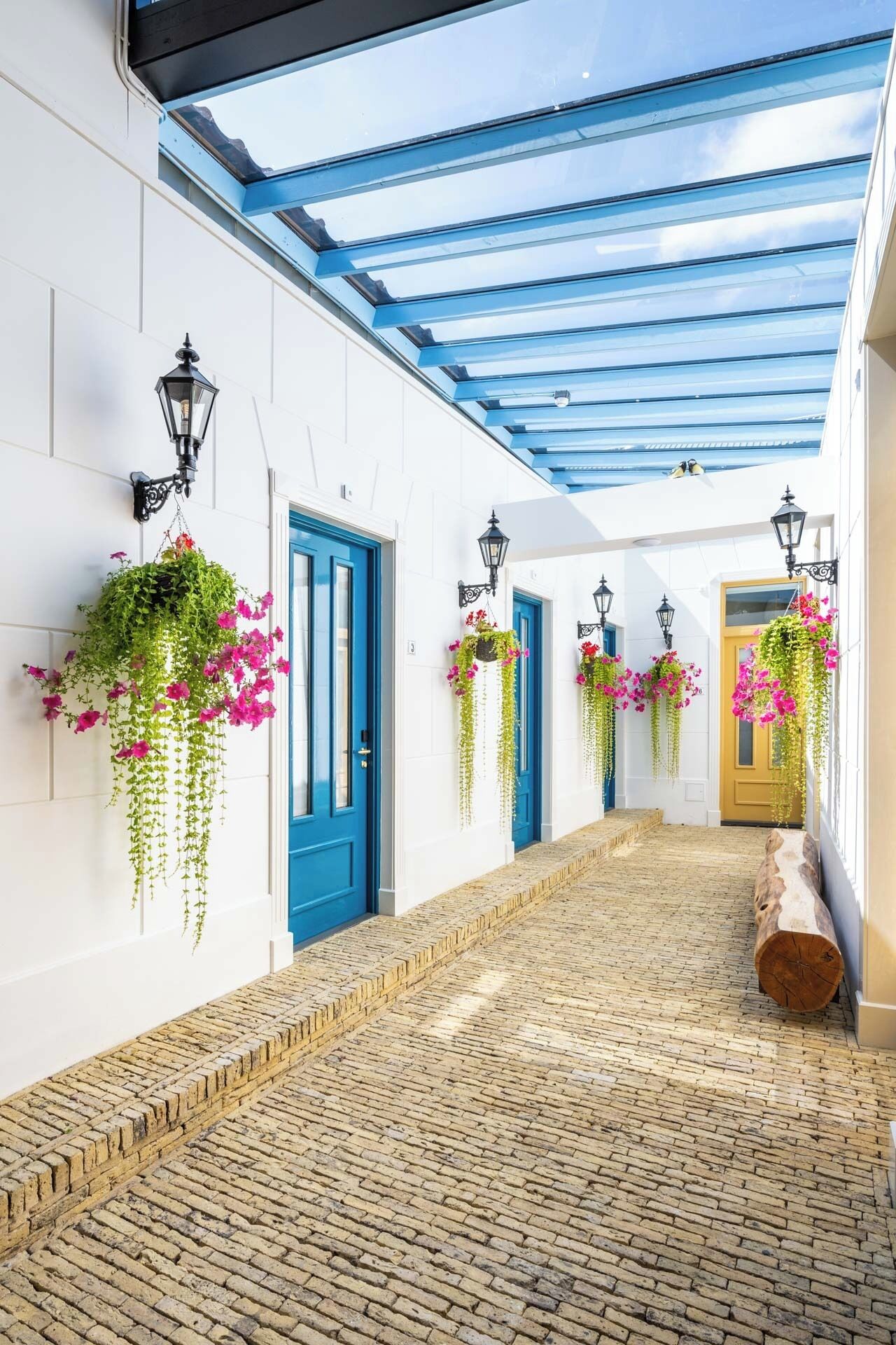 A corridor with a cobblestone floor, white walls, blue doors, and hanging flower baskets.
