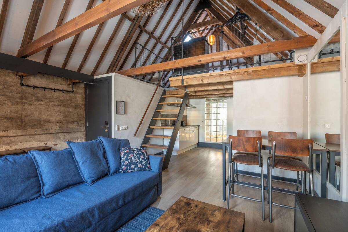 A loft-style room with exposed wooden beams, a blue sofa, and a dining area with metal chairs.