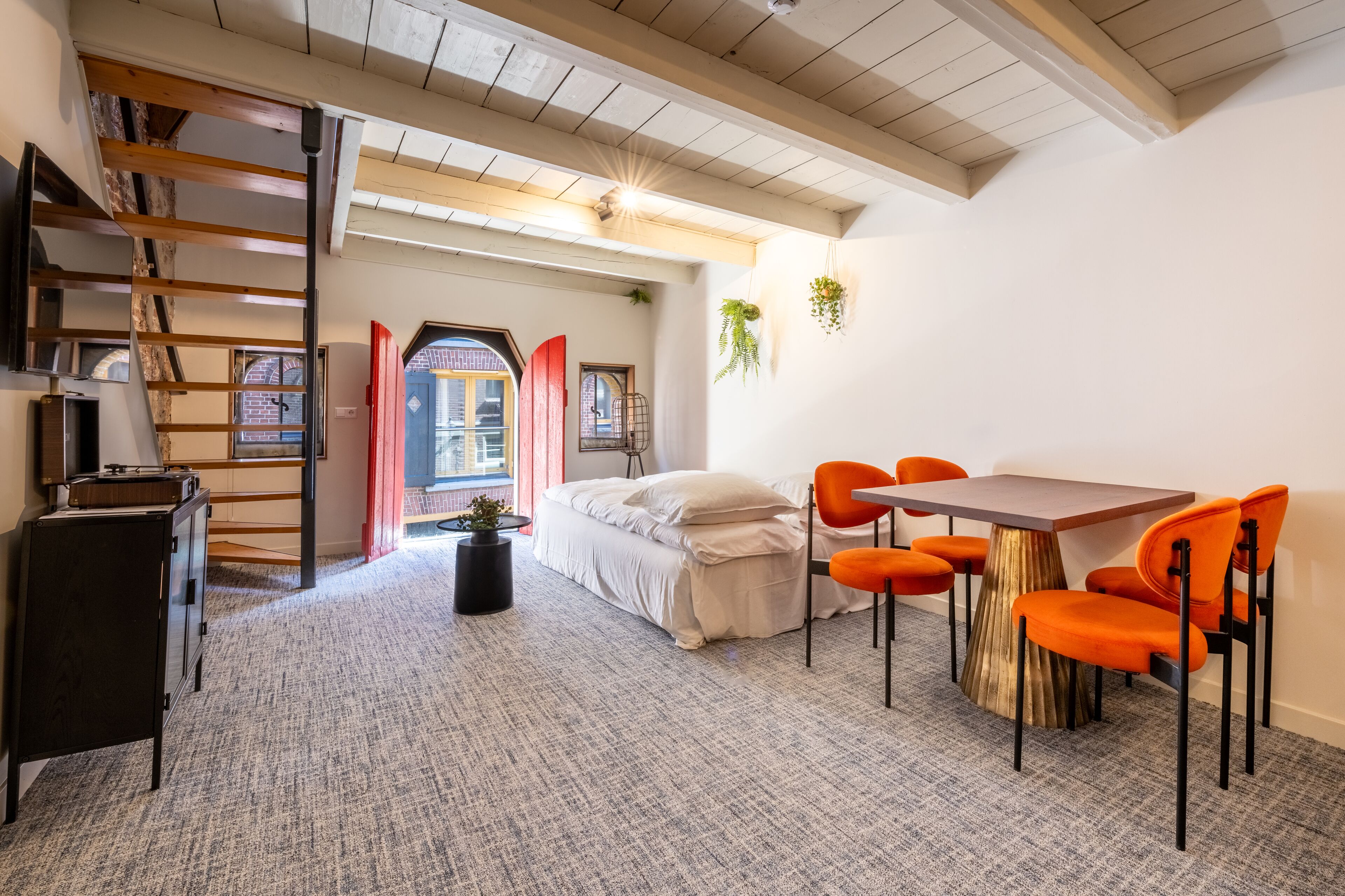A modern bedroom with a loft-style bed, a dining area with orange chairs, and a small table.