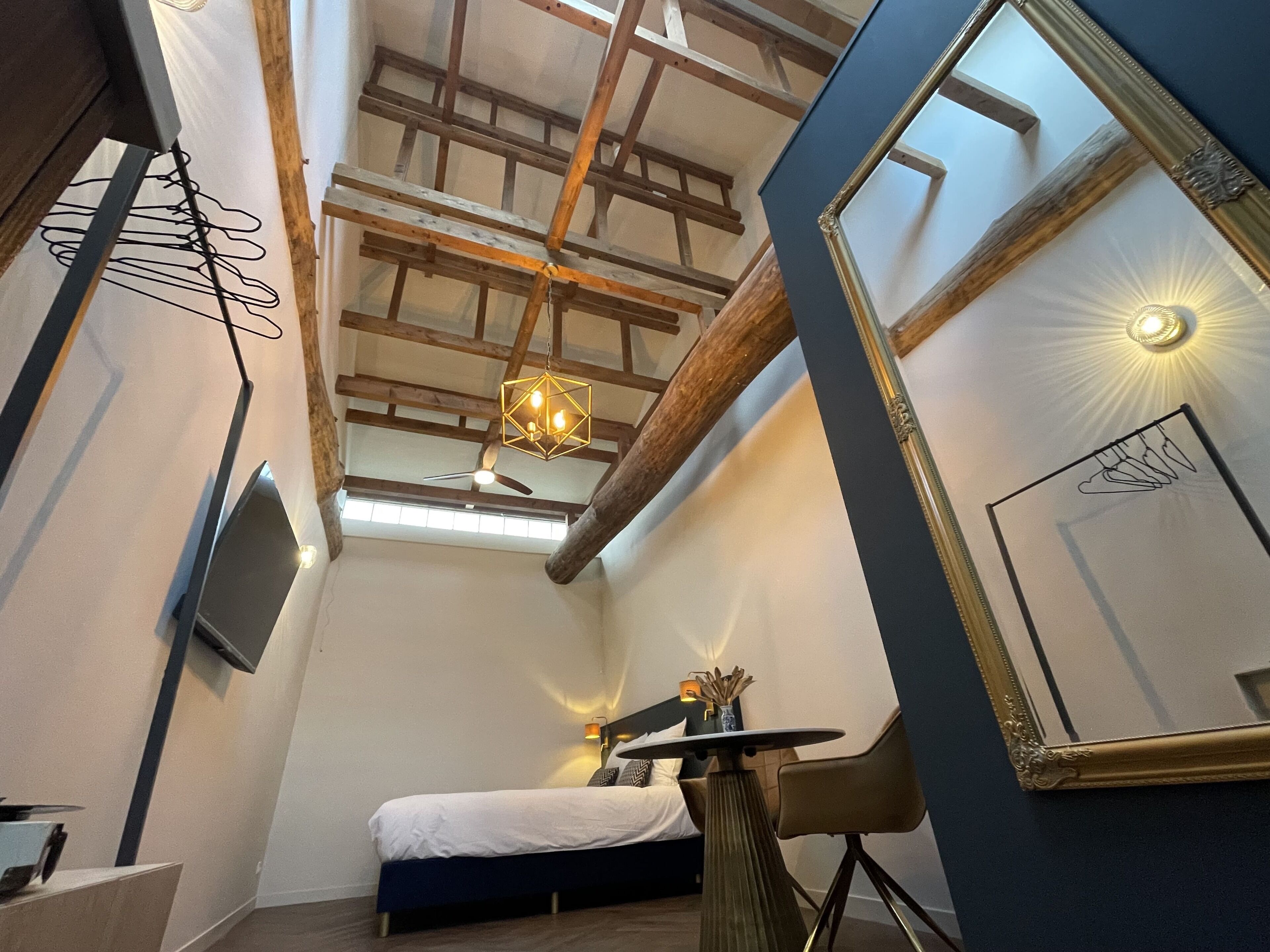 A narrow room with a bed, a table, and a chair. There is a ceiling fan, a hanging light fixture, and a large mirror.