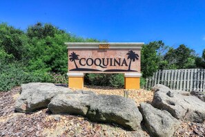 A sign with the name 'COQUINA' and palm tree designs, set against a backdrop of greenery and a wooden fence.