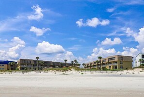 A beachfront hotel with multiple buildings, a sandy beach, and a clear blue sky with clouds.