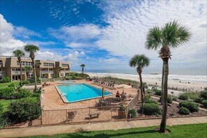 A pool area with a view of the beach, surrounded by palm trees and a building with multiple balconies.