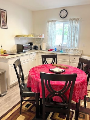 Cocina compacta con armarios blancos, microondas y cafetera. Hay una mesa de comedor con cuatro sillas, cubierta con un mantel rojo. Se ve un reloj en la pared y una ventana con cortinas.