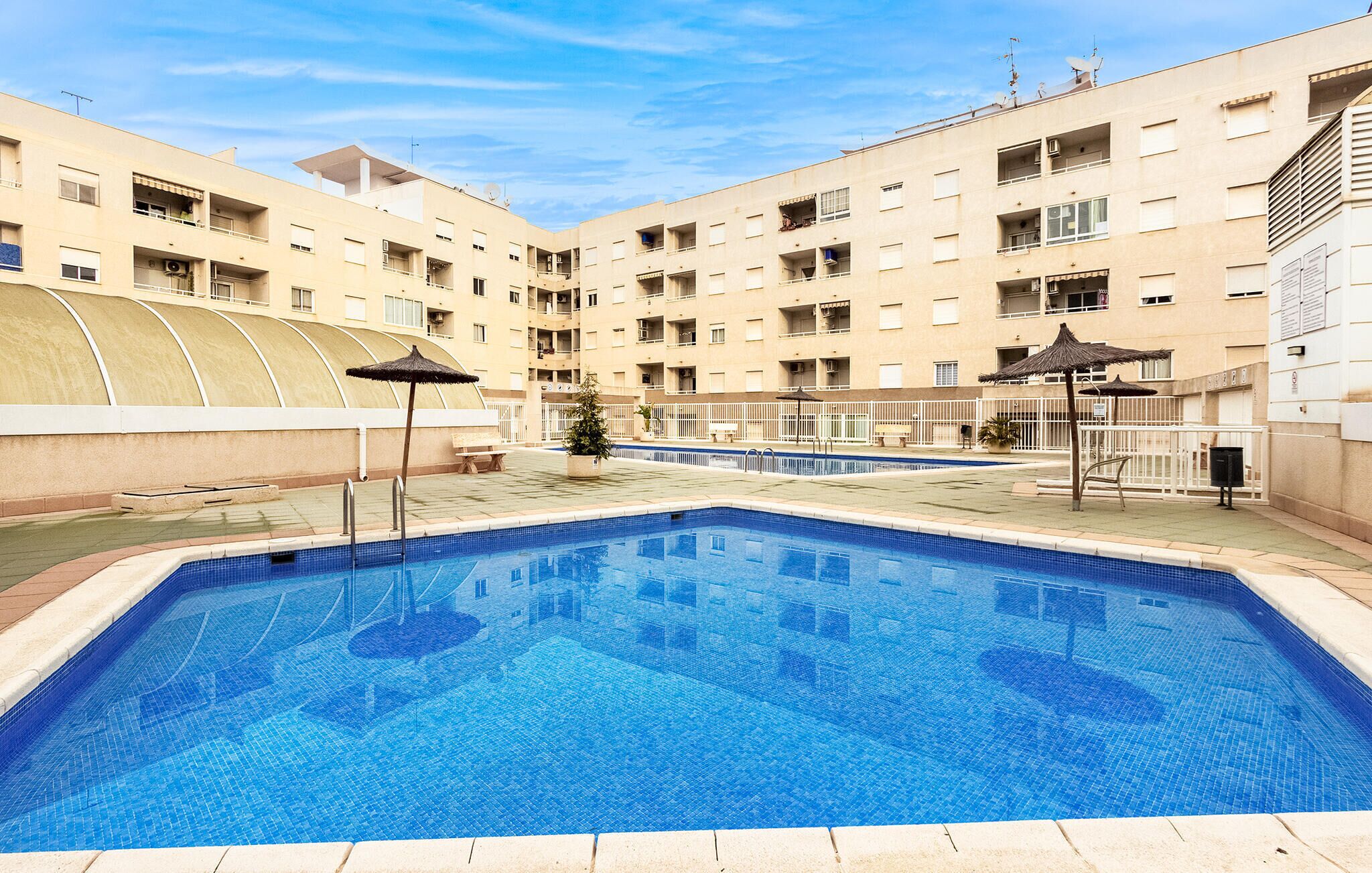 A swimming pool with a tiled deck, surrounded by a multi-story building with balconies and a covered area.