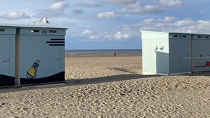 Cabañas de playa con los números 25 y 26, una gaviota encima, una playa de arena y el mar al fondo.