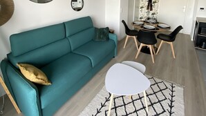 Un sofá color turquesa con un cojín dorado, una mesa de centro blanca y una alfombra estampada en un salón moderno con un comedor al fondo.