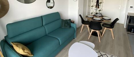 A teal sofa with a gold pillow, a white coffee table, and a patterned rug in a modern living room with a dining area in the background.