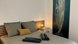 Una cama bien hecha con una colcha gris, dos almohadas y toallas enrolladas. Una mesita de noche con lámpara está al lado de la cama.